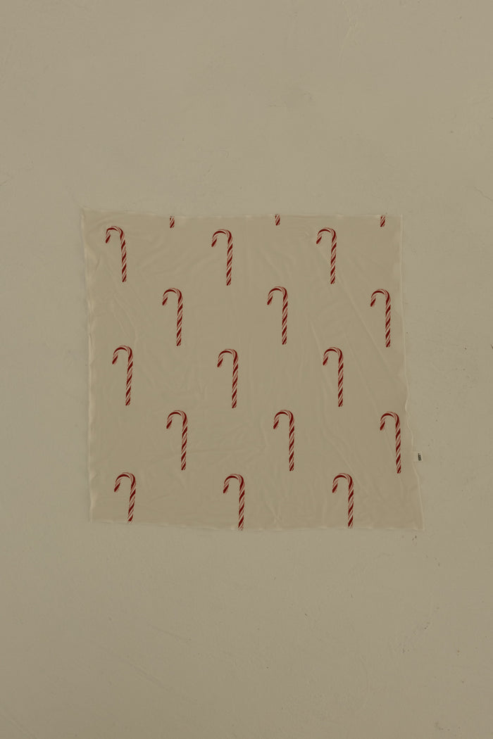 The Bamboo Bandana | Candy Cane by forever french baby, featuring a repeating candy cane print, is taped to a plain beige wall.