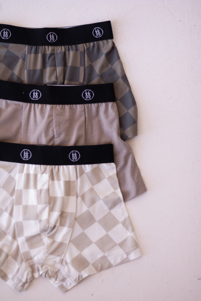The Bamboo Boxers (3 Pack) by forever french baby features checkerboard and faded brown patterns with black waistbands—soft, hypo-allergenic underwear laid flat on a light surface for stylish comfort.