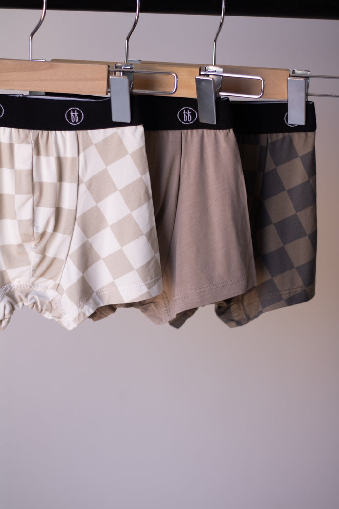 Three pairs of forever french baby Bamboo Boxers (3 Pack) hang on wooden hangers, featuring checkerboard and faded brown checkerboard designs. Enjoy stylish, hypo-allergenic comfort in these premium men’s underwear.