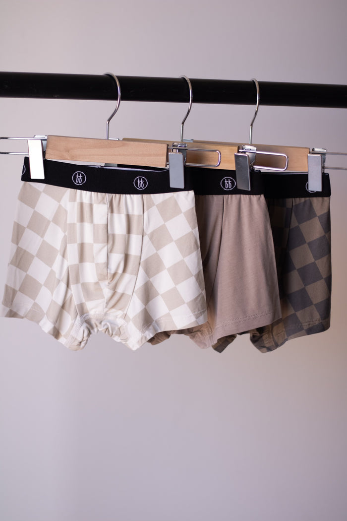 Three pairs of Bamboo Boxers (3 Pack) by forever french baby—Checkerboard and Faded Brown Checkerboard—hang from wooden clip hangers on a black rack. Enjoy hypo-allergenic comfort in stylish, neutral tones.