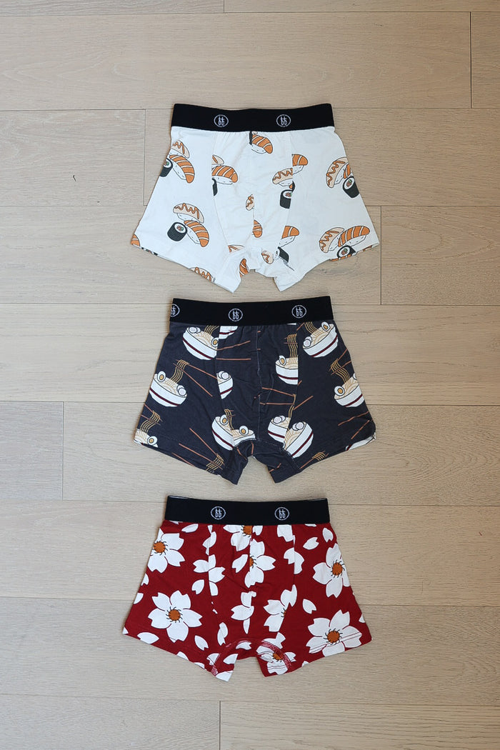 The forever french baby Bamboo Boxers (3 Pack) set features boys’ boxers in fun prints: sushi, ramen bowls, and red with white cherry blossoms, all laid out on a wooden floor.