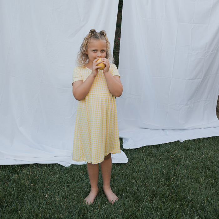 A young girl in the forever french baby Bamboo T-Shirt Dress | Citron stands barefoot on grass, holding a yellow fruit. White fabric behind her creates a soft, hypo-allergenic backdrop.