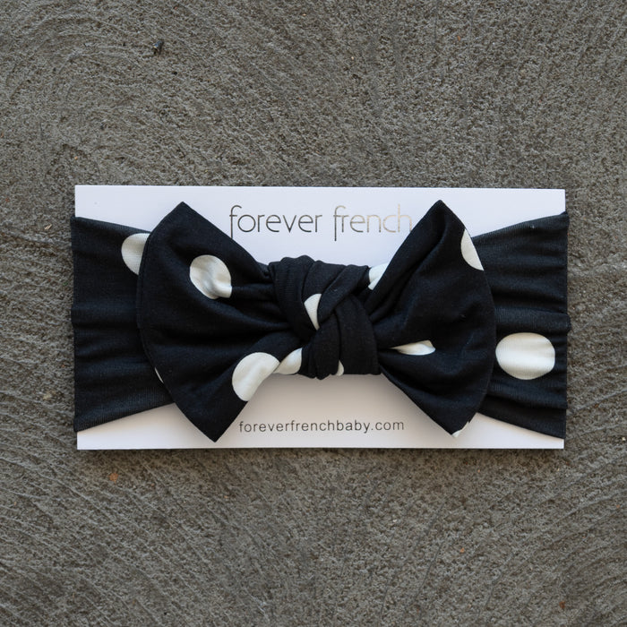 Showcased on a textured gray surface, the Bamboo Head Wrap | B&W Polka Dot from forever french baby features bold white polka dots on black and is styled with an adorable bow, presented on a branded “forever french” card.