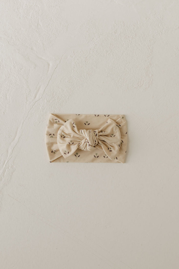 The Flora Bamboo Head Wrap from forever french baby, in beige with a central bow showcasing a subtle pattern of small black dots, lies elegantly on a light, textured surface.