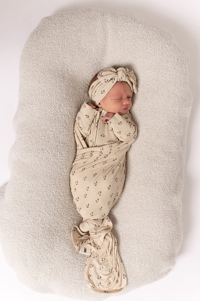 A newborn is swaddled in a beige blanket with small patterns, wearing the forever french baby Bamboo Head Wrap | Flora, lying on a soft light gray cushion against a plain white background.
