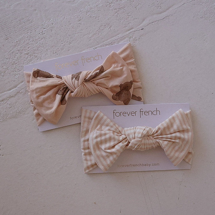 Two forever french baby “Bamboo Head Wrap | Lottie” headbands on branded cards: one with a teddy bear print, the other in beige and white gingham, both displayed on a light surface.