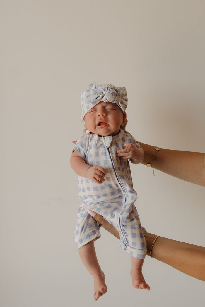 A newborn baby wearing the Forever French Baby Bamboo Head Wrap | Marilyn is being held up by an adult’s hands against a plain light background. Styled in signature forever french baby fashion, the baby has their eyes closed and appears to be crying.