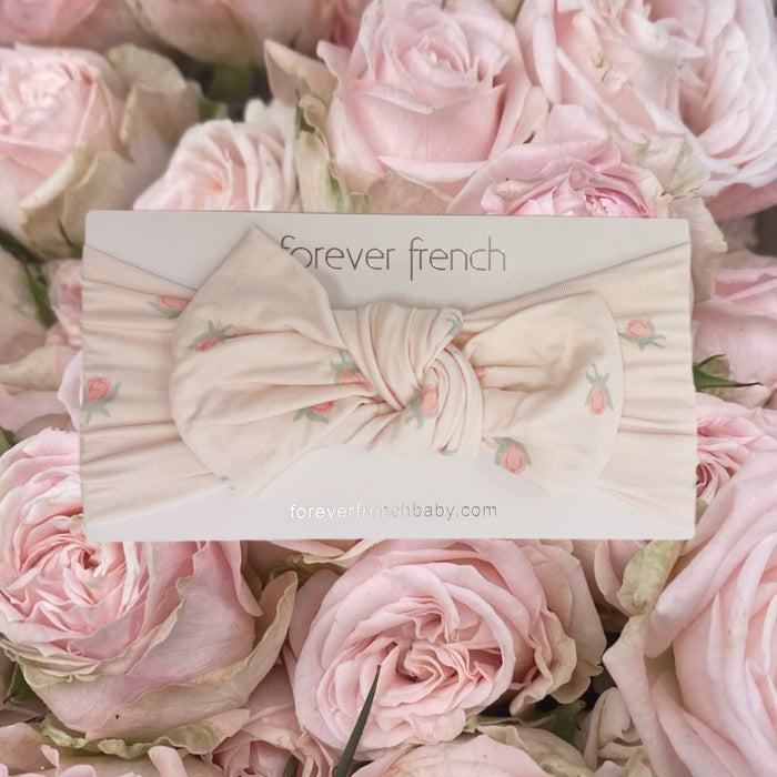 A cream-colored Bamboo Head Wrap | Rosie from forever french baby, featuring small pink floral prints, is displayed on a “forever french” card and surrounded by light pink roses. This sweet, adjustable head wrap is perfect for delicate little heads and adds a charming touch to any outfit.