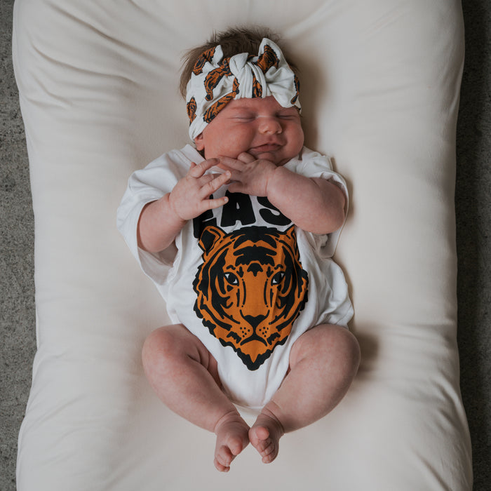 A baby wearing the forever french baby Bamboo Head Wrap | Tigers and a white onesie with a tiger face graphic smiles with eyes closed, lying on a soft white cushion.
