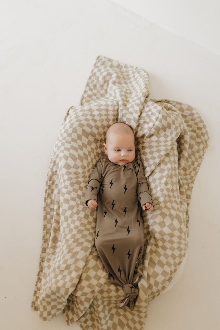 A baby rests on a soft checkered blanket, wrapped in the forever french baby Bamboo Knotted Gown featuring brown fabric adorned with black lightning bolts. The hypoallergenic materials enhance the calm and comfort of the scene.