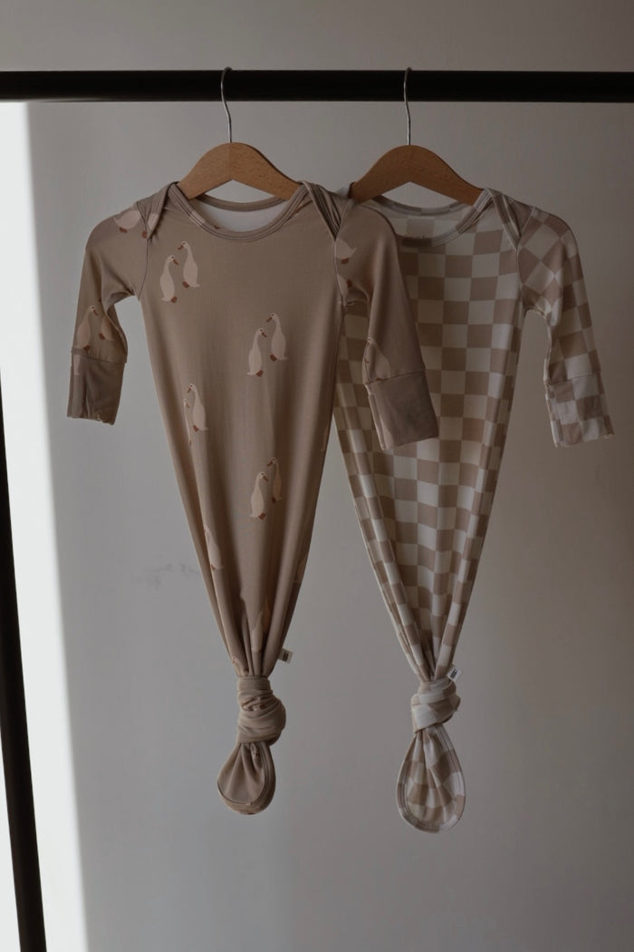 Two hypoallergenic Bamboo Knotted Gowns from forever french baby hang on wooden hangers: one features penguin prints, and the other showcases a beige and white checkerboard pattern, both displayed against a neutral background.