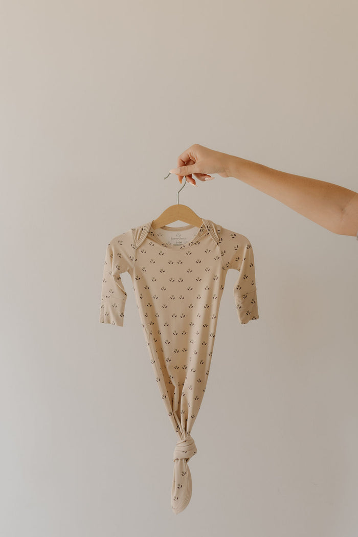 A hand holds a bamboo "Flora" knotted gown from forever french baby, designed for newborns. It's beige with subtle black motifs and hangs on a wooden hanger against a light-colored wall. Hypoallergenic and essential, it's crafted from eco-friendly materials.