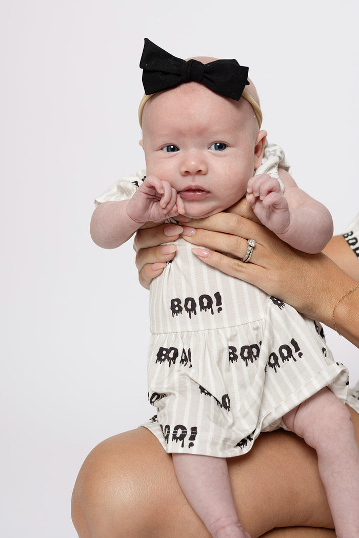A baby wears the forever french baby Bamboo Ruffle Romper | Boo! in cream with a black "BOO!" print and matching black bow headband, offering hypoallergenic comfort as the baby looks forward with a neutral expression.