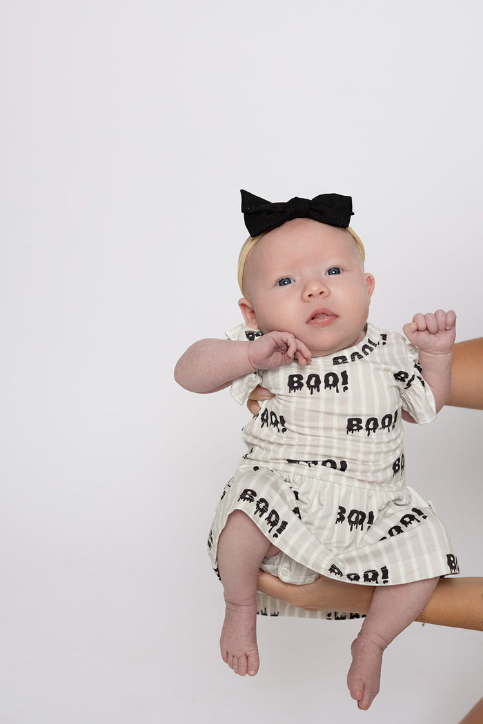 A baby in the forever french baby Bamboo Ruffle Romper | Boo! with black patterns and a black bow headband is held up by an adult hand against a plain light background.