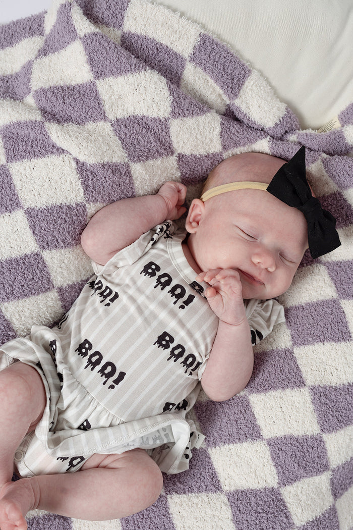 A baby in the forever french baby Bamboo Ruffle Romper | Boo! and a black bow headband sleeps peacefully on a purple and white checkered blanket, one hand near their face.