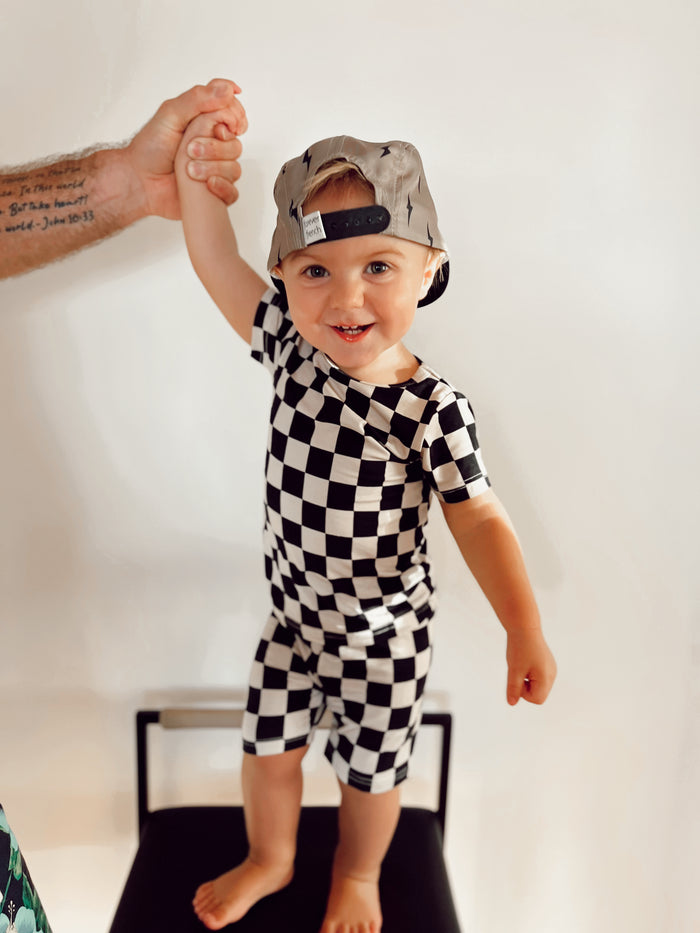 A cheerful toddler stands on a chair holding an adult's hand, dressed in the Bamboo Short Set in Black Checkerboard by forever french baby. The child wears a gray baseball cap backwards, set against a plain, light-colored background.