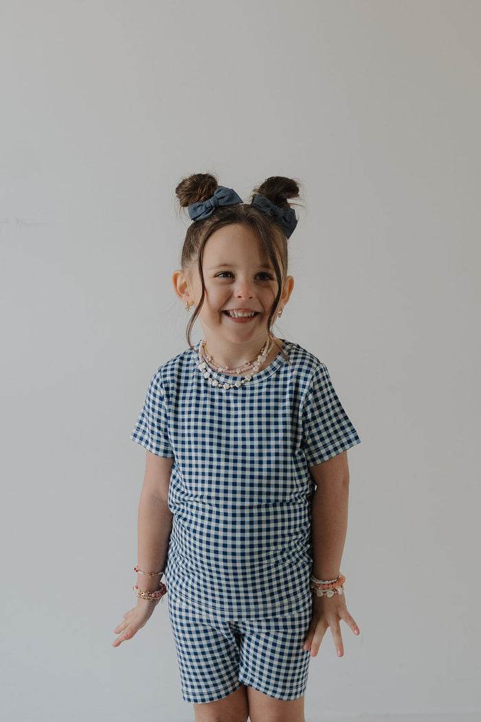 A young child smiles against a plain light background, wearing the forever french baby Bamboo Short Set in Blue Gingham, complete with matching hair bows, beaded bracelets, and a beaded necklace.