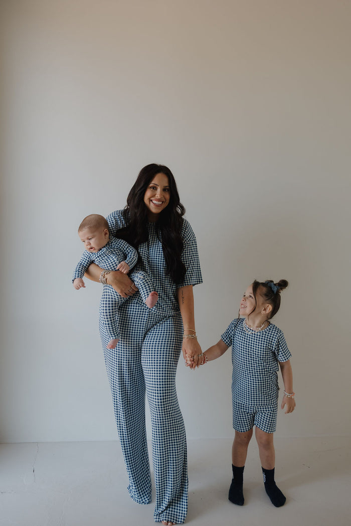 A woman with a baby and a young girl, all wearing forever french baby Bamboo Short Set in Blue Gingham, stand together against a plain light-colored wall.