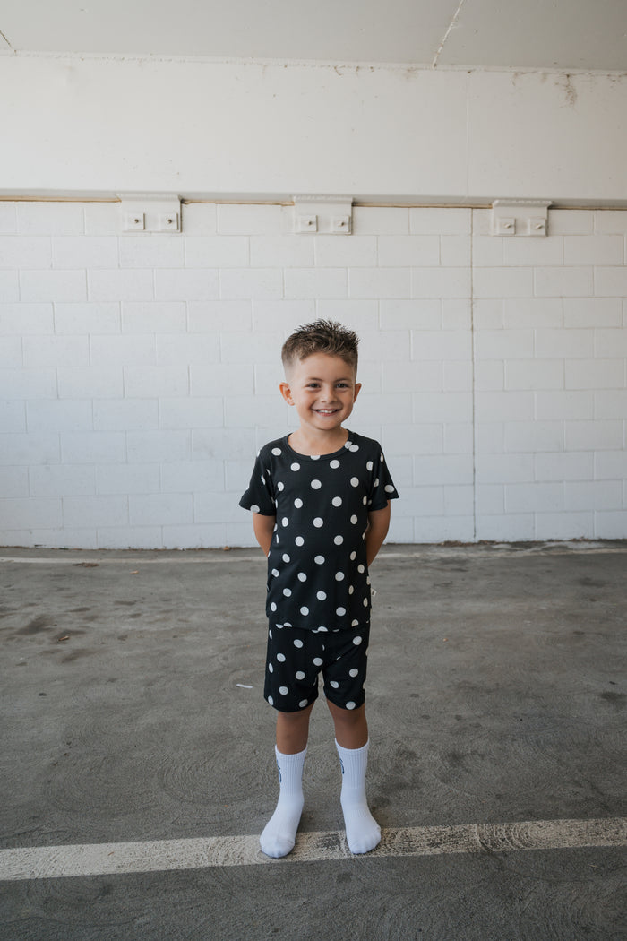 A smiling young boy stands with his hands behind his back in a parking garage, wearing the forever french baby Bamboo Short Set in B&W Polka Dot—a breathable black shirt and shorts adorned with white polka dots.