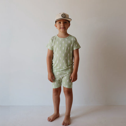 A young child stands barefoot against a white background, wearing the forever french baby Bamboo Short Set | Flash—light green matching t-shirt and shorts with white lightning bolts—hypo-allergenic, stylish, and comfy, plus a light baseball cap.