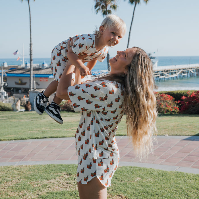 A woman with long blonde hair lifts a smiling toddler in the air. Both wear matching forever french baby Bamboo Short Sets | Fries & Cola, featuring a brown pattern, outdoors by grass and the ocean on a sunny day.
