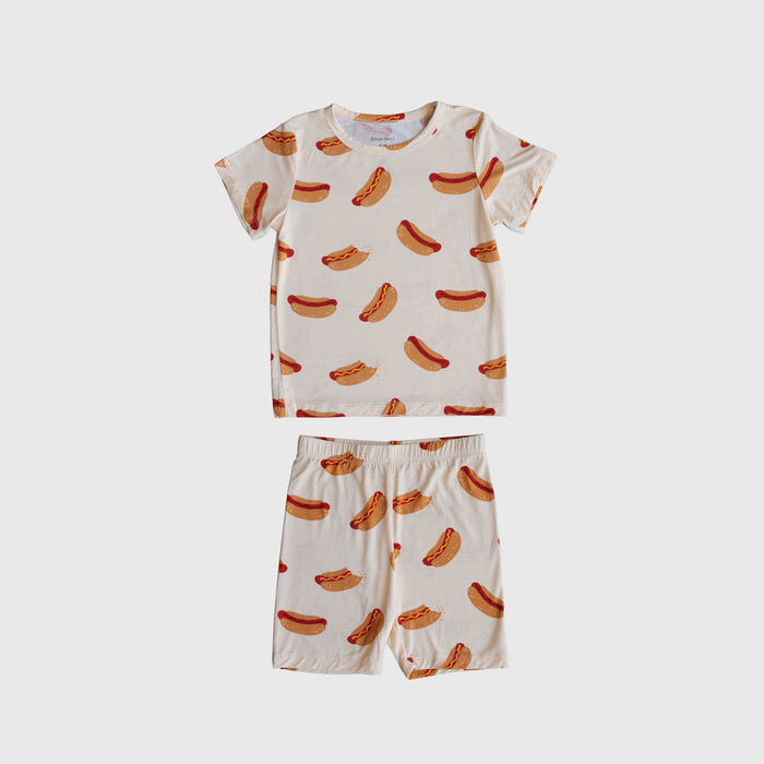 The Bamboo Short Set | Hot Dogs by forever french baby features a cream short-sleeve shirt and shorts with an all-over hot dog print, made from breathable sleepwear fabric and shown on a plain white background.
