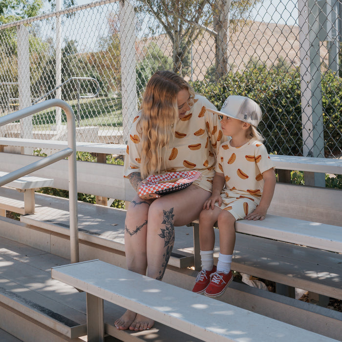 A woman and young child wearing matching forever french baby Bamboo Short Sets in the Hot Dogs print smile at each other on outdoor bleachers. The woman has long hair and tattoos, while the child wears a cap and red shoes.