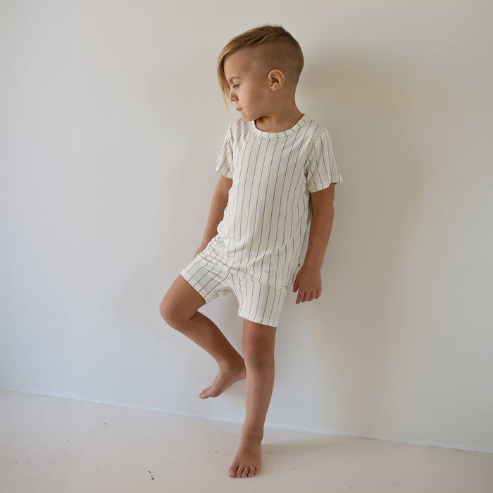 A young child with short, side-shaved hair stands barefoot against a white wall, wearing the forever french baby Bamboo Short Set | Jack—a breathable cream-colored kids’ outfit with thin black vertical stripes.