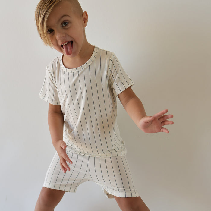 A young child with straight blonde hair and side-swept bangs poses playfully, wearing the Bamboo Short Set | Jack by forever french baby—a cream-colored, short-sleeve top and striped shorts—against a plain background.