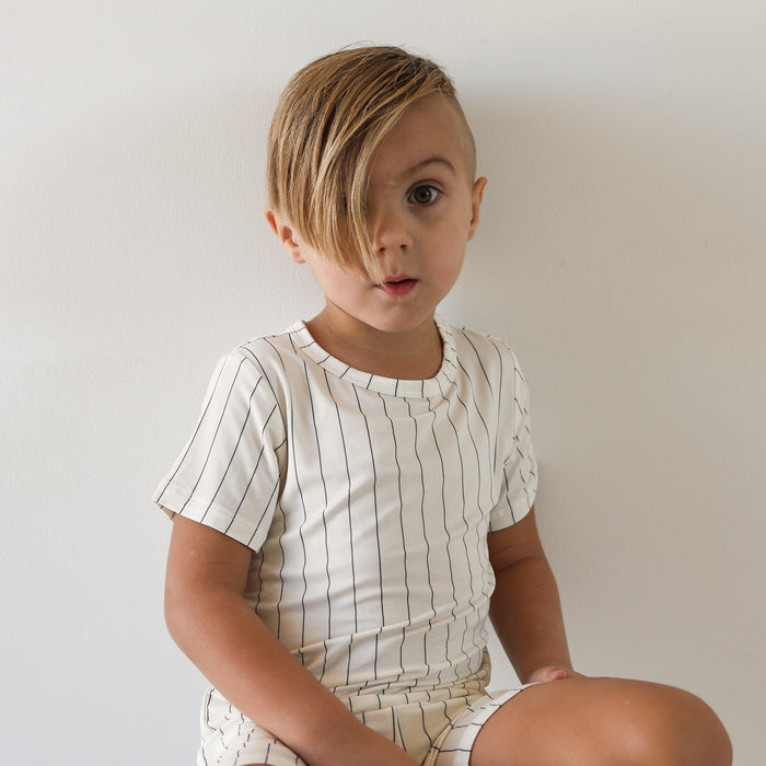 A young child with light brown side-swept hair wears forever french baby's Bamboo Short Set | Jack—a breathable white shirt with thin black stripes—while sitting against a plain white background and looking surprised.