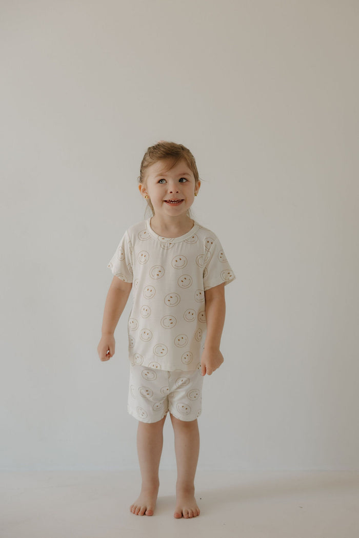 A young child with light brown hair smiles slightly, wearing the forever french baby Bamboo Short Set | Just Smile—cream-colored bamboo pajamas with a smiley face pattern—standing barefoot against a plain background. The breathable set keeps them comfortable.