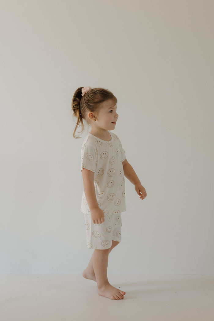 A young child with light brown hair tied back stands barefoot indoors against a plain wall, wearing the soft, hypo-allergenic Bamboo Short Set | Just Smile by forever french baby, featuring a cheerful smiley face pattern.