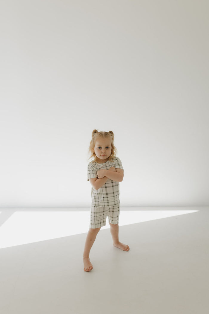 A young child with blonde hair, wearing the Bamboo Short Set in Picnic Plaid by forever french baby, stands barefoot on a white surface. With arms folded confidently, soft shadows from natural light play against the white wall backdrop. The outfit is made of breathable fabric.