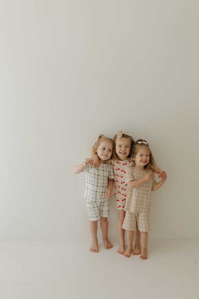 Three smiling young children stand barefoot against a plain background, embracing while wearing matching Bamboo Short Sets in Picnic Plaid by forever french baby. The breathable fabric and charming checkered and heart patterns add to the delightful scene.