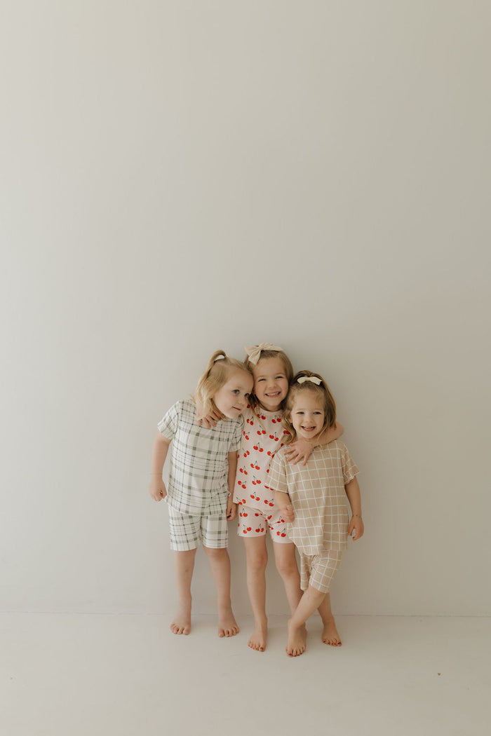 Three barefoot children stand against a light wall in forever french baby pajamas. They wear the Bamboo Short Set in Picnic Plaid, cherries, and grid designs. Their smiles reflect the comfort of their breathable outfits as they lean toward each other.