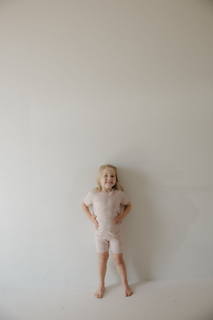 A young child with blonde hair smiles barefoot against a light wall, wearing the breathable Kendy 3 Bamboo Short Set | Ruby—light-colored, short-sleeved pajamas featuring subtle patterns.