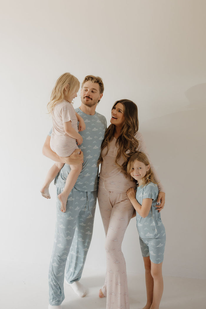 A smiling family of four wears matching Kendy 3 Bamboo Short Set | Duke pajamas. Barefoot, the father holds a young girl while the mother hugs their older daughter, all posing together against a plain background.