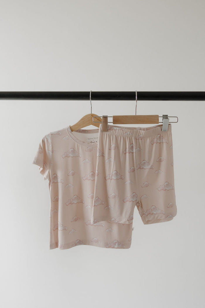 The Kendy 3 Bamboo Short Set | Ruby, a breathable light pink pajama set with a cloud pattern, hangs on wooden hangers and is displayed on a black rack against a plain white background.