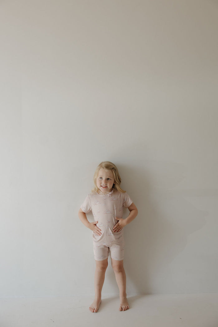 A young child with shoulder-length blonde hair stands barefoot against a light wall, smiling hands on hips while wearing the Kendy 3 Bamboo Short Set | Ruby—a breathable, light pink matching t-shirt and shorts set.