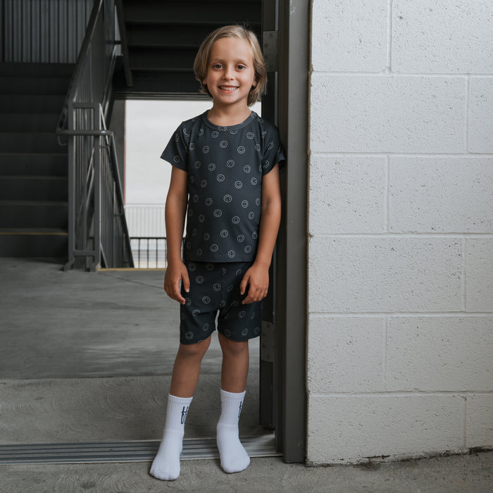 A young blonde child smiles indoors by a white brick wall, wearing forever french baby’s Bamboo Short Set | Smiley Ribbed pajamas and white socks. Stairs and a metal railing in the background highlight these cozy, hypoallergenic PJs.