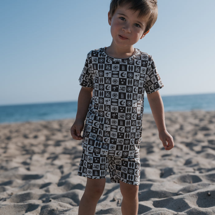 A young child wearing forever french baby’s Bamboo Short Set | Summer Check stands on a sandy beach, their comfy, hypoallergenic pajamas contrasting beautifully with the sea and clear blue sky in the background.