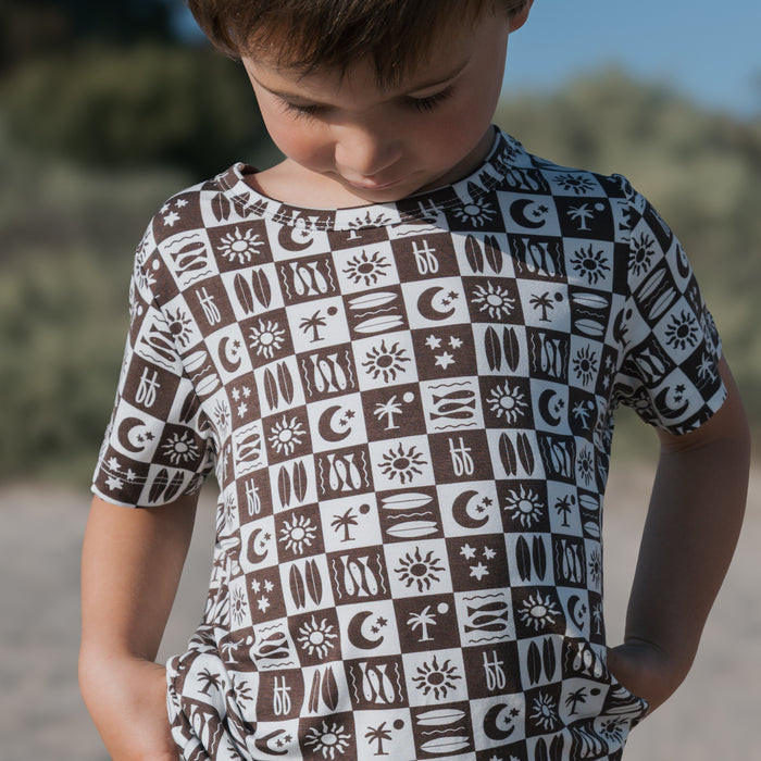 A young child stands outdoors in the Bamboo Short Set | Summer Check by forever french baby, looking down with hands in pockets. The breathable outfit features a summery check pattern; blurred greenery and blue sky fill the background.