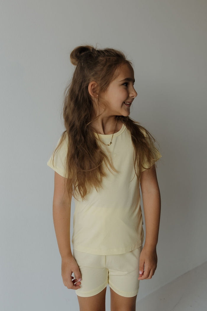 A young girl with long brown hair in a half-up style is wearing the forever french baby Bamboo Short Set | Sunny—a light yellow, breathable and comfy t-shirt with matching shorts—standing against a plain, light background and smiling as she looks to the side.