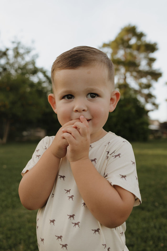 A young child smiles softly outdoors on grass, wearing the forever french baby Bamboo Short Set in Wild Horses—a light-colored shirt with animal prints. Trees blur in the background as the child holds their hands near their mouth.