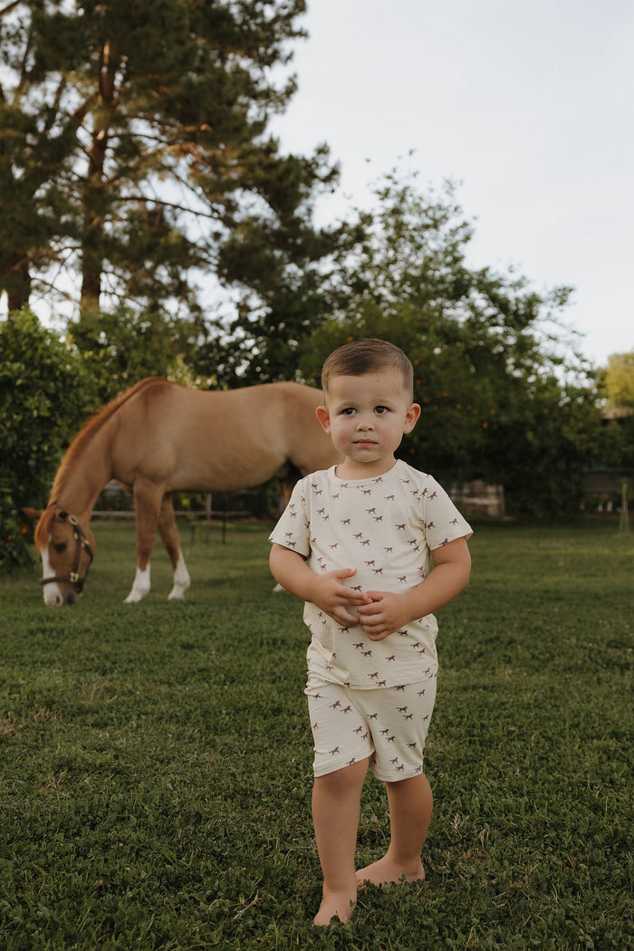 A young child wears the Bamboo Short Set | Wild Horses by forever french baby, standing barefoot on grass near a grazing light brown horse, surrounded by trees and greenery in an outdoor setting.