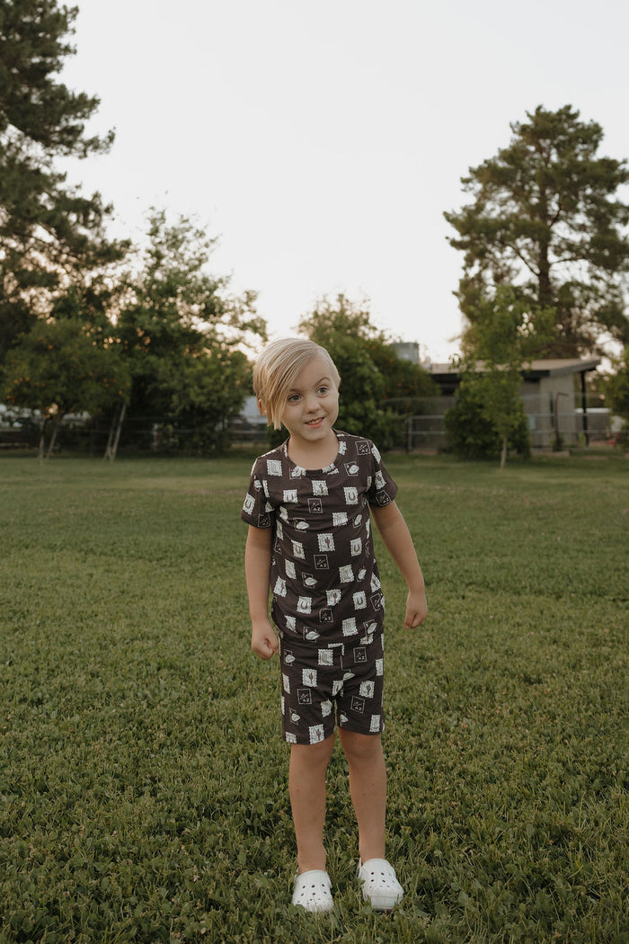 A young child with light hair stands on a grassy lawn, wearing the Bamboo Short Set by forever french baby. White shoes complete the look. In the background are trees and a building under a clear sky.