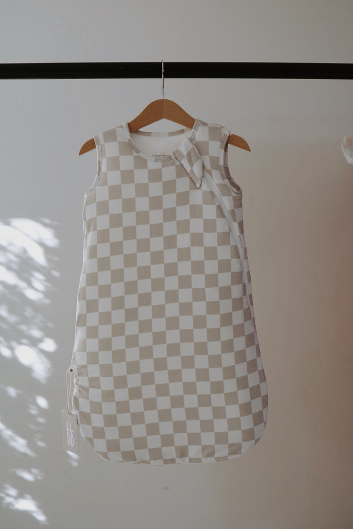The Bamboo Sleep Sack | Checkerboard by forever french baby, in beige and white checks, hangs on a wooden hanger against a plain background with soft sunlight casting shadows on the left.