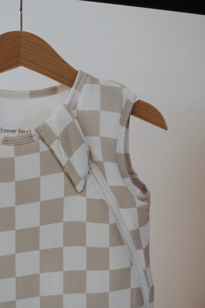 The forever french baby Bamboo Sleep Sack | Checkerboard, in beige and white checks, hangs on a wooden hanger and features a zipper with a snap button on the shoulder.