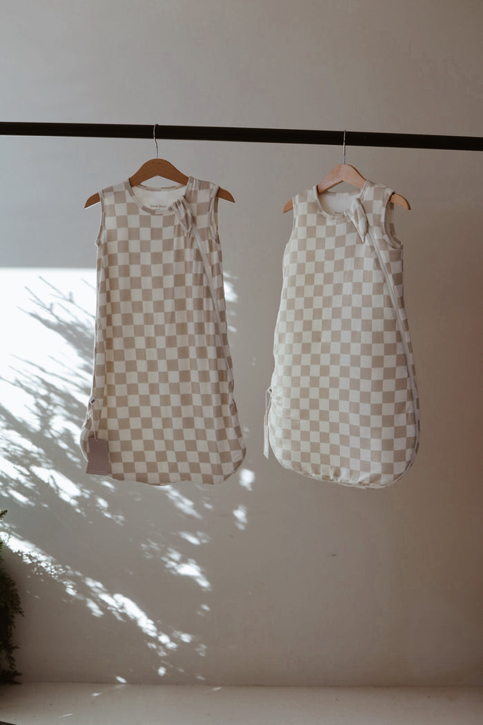 Two forever french baby Bamboo Sleep Sack | Checkerboard in beige and white hang on wooden hangers from a rod, with sunlight casting plant shadows on the wall behind them.
