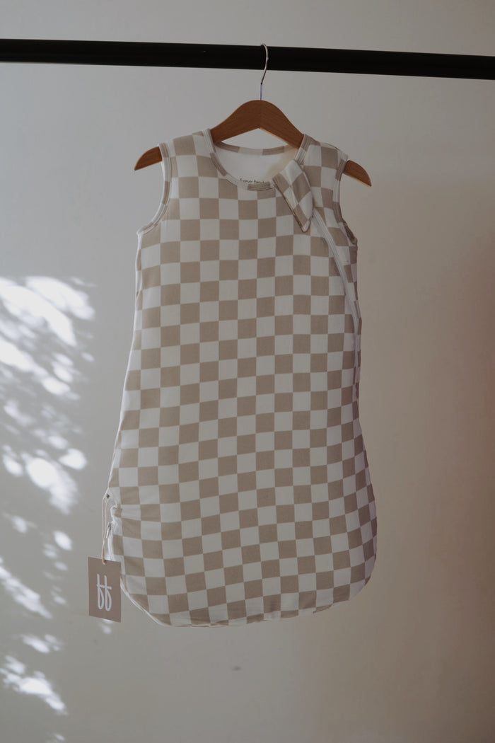 The Bamboo Sleep Sack | Checkerboard from forever french baby, featuring a beige and white checkerboard print, hangs on a wooden hanger as sunlight casts soft shadows. A "bb" tag accents this sleeveless, cozy wearable blanket.